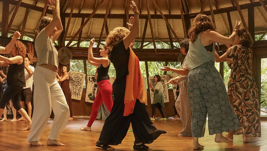 Gente bailando en una estructura con techo de madera, con los brazos en alto. La luz del sol entra a raudales.