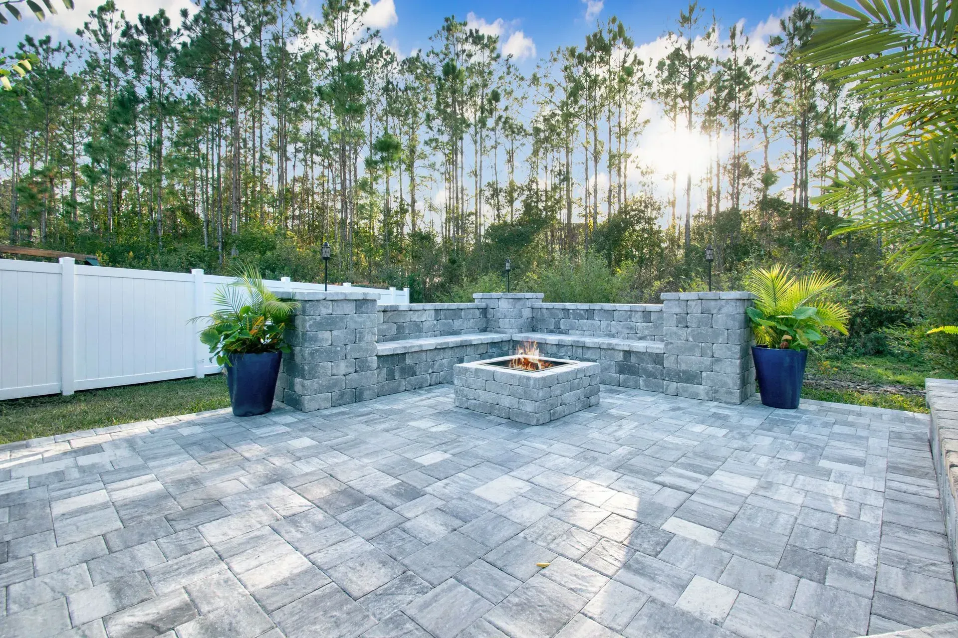 Paver patio with built-in stone bench seating, a square fire pit, and tropical plants in blue pots, 