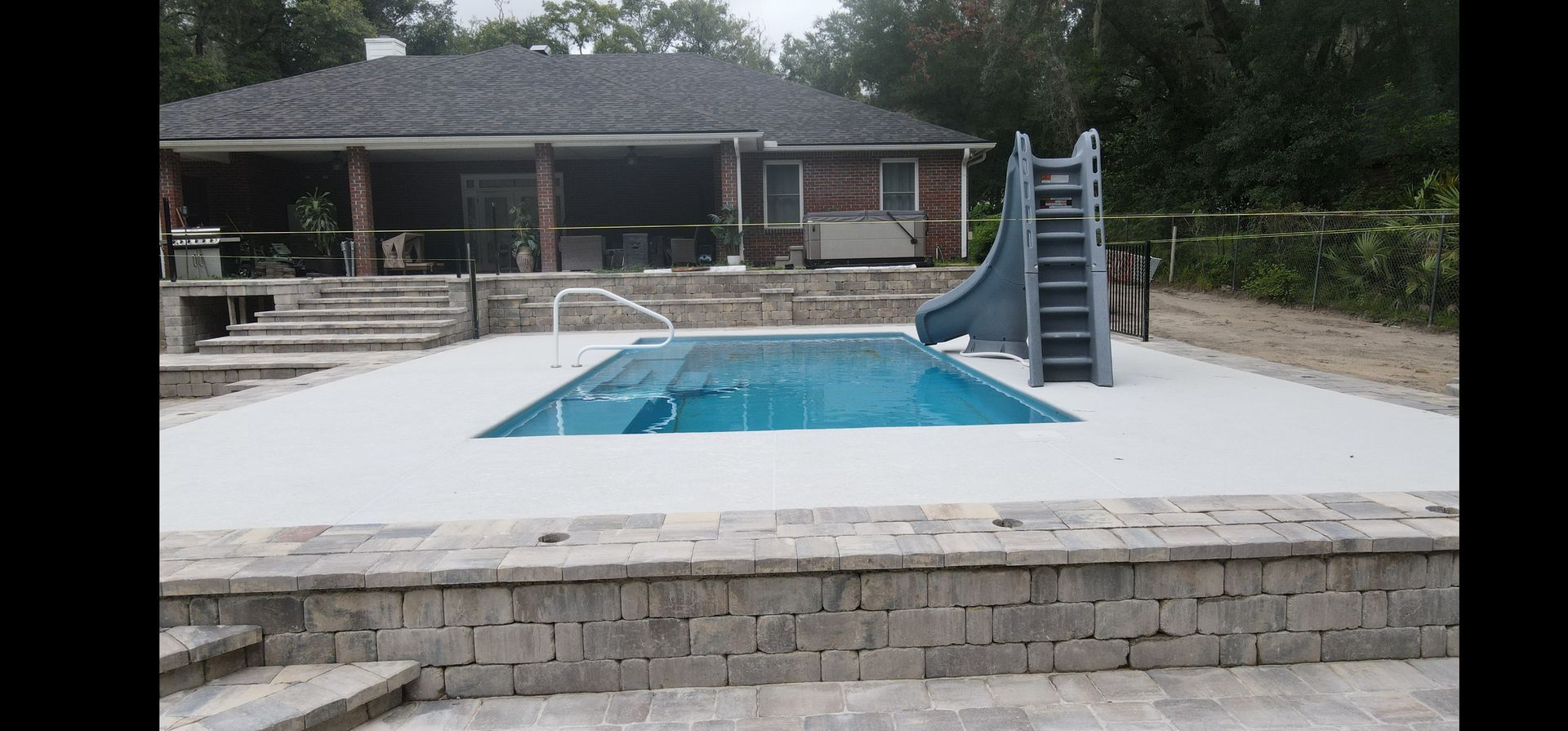 A large swimming pool with a slide and pavers around it in front of a house.
