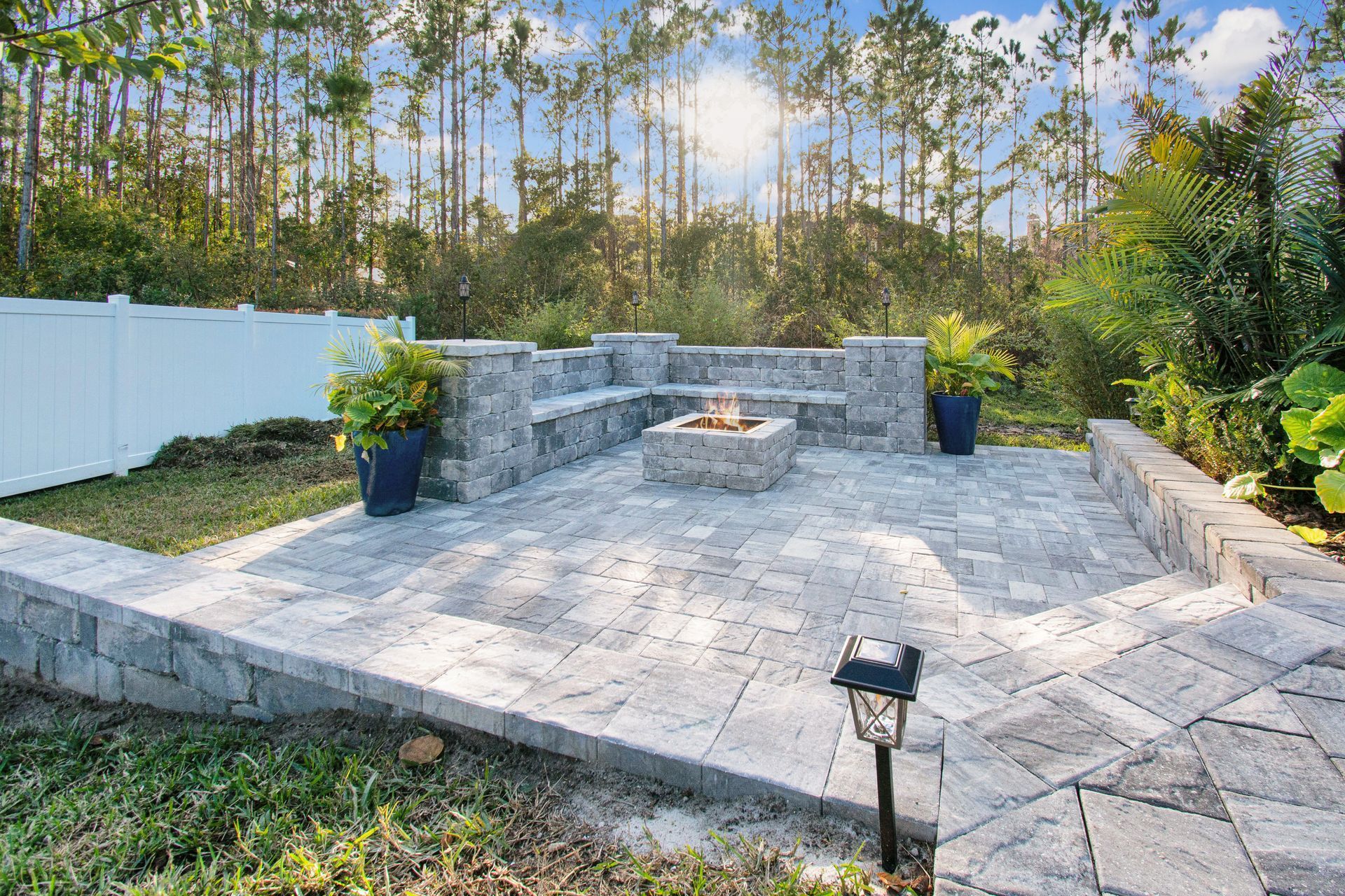 There is a fire pit in the middle of the paver patio with steps and a retaining wall