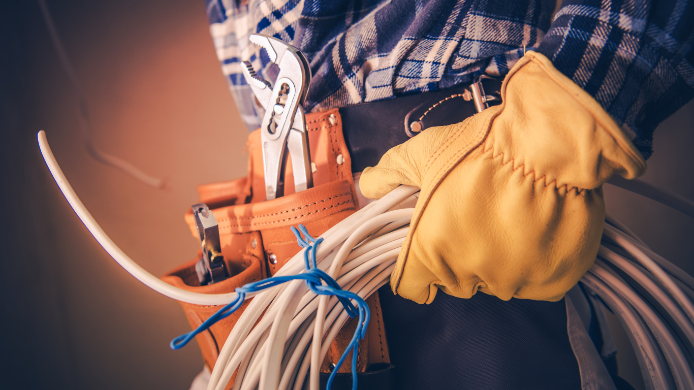 Electrician with a tool belt, holding a bundle of white wires and a wrench, wearing yellow gloves.