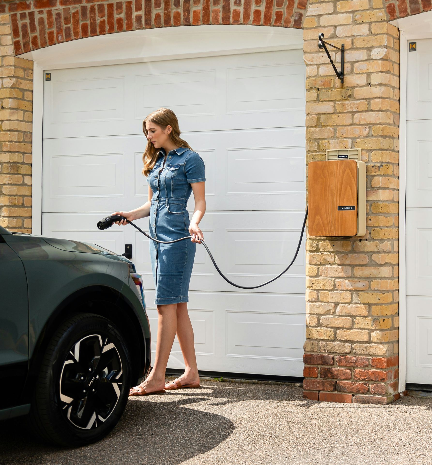 Woman charging an electric car in front of a garage. She is wearing a denim dress. The charger is mounted on a brick wall.