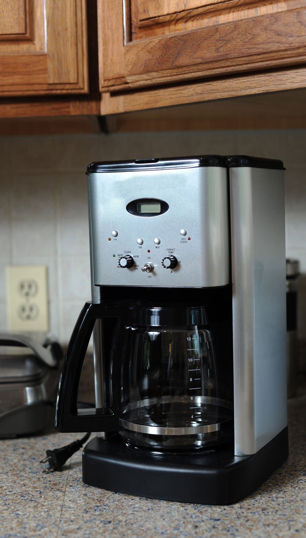 Silver coffee maker on a countertop, with a black carafe. Electrical outlet and toaster oven in background.