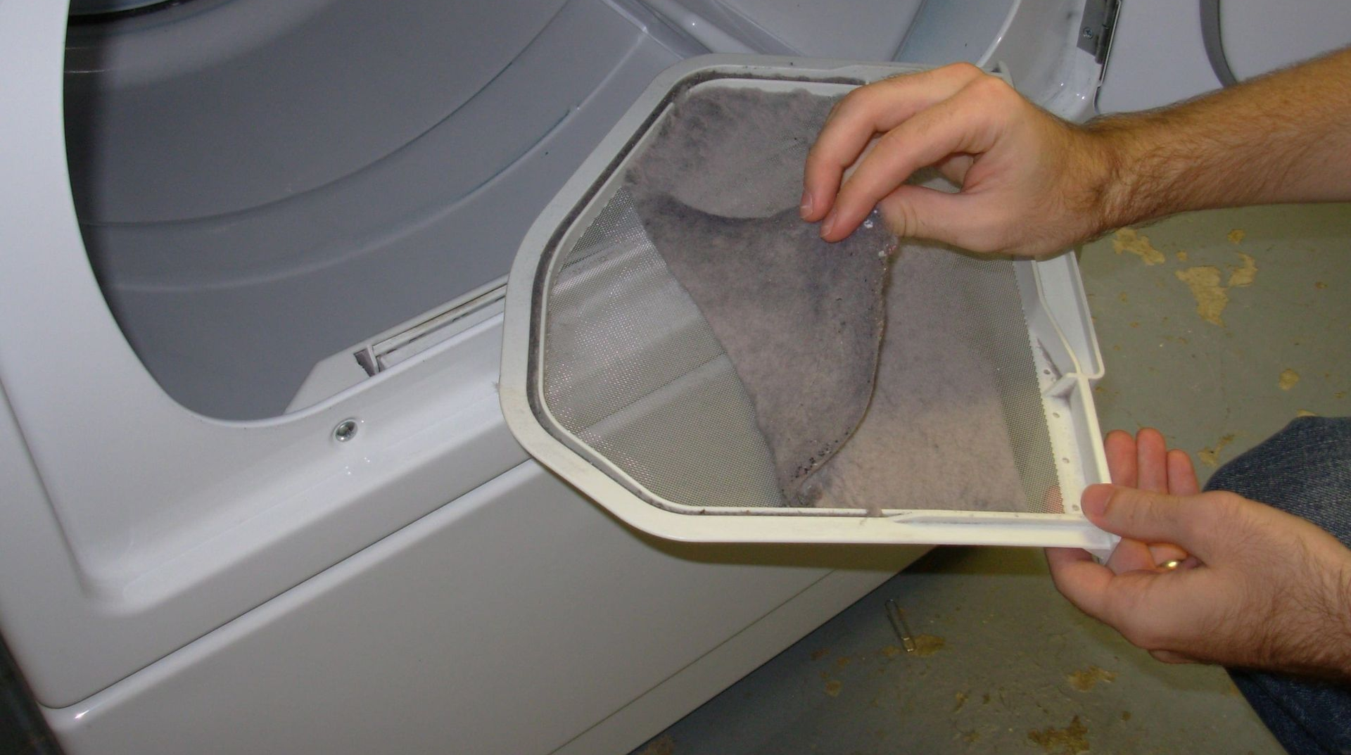 Person removing lint from a dryer filter. White dryer in a utility room.
