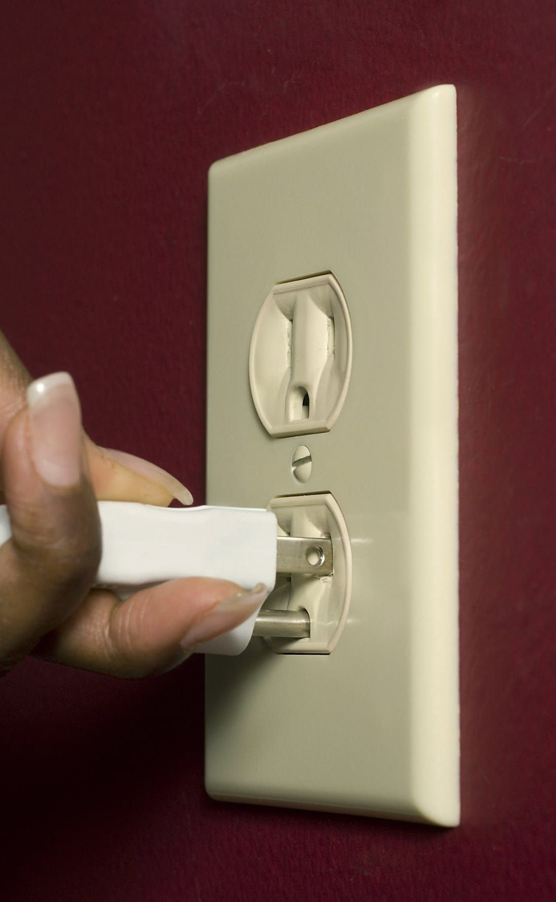 Hand plugging a white electrical plug into an outlet on a maroon wall.