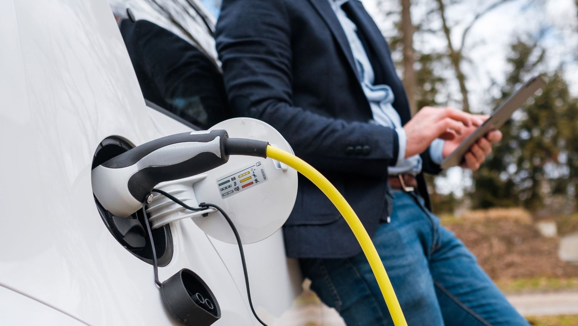 Man in a suit leaning against a white electric car while using a tablet, charging cable plugged in.