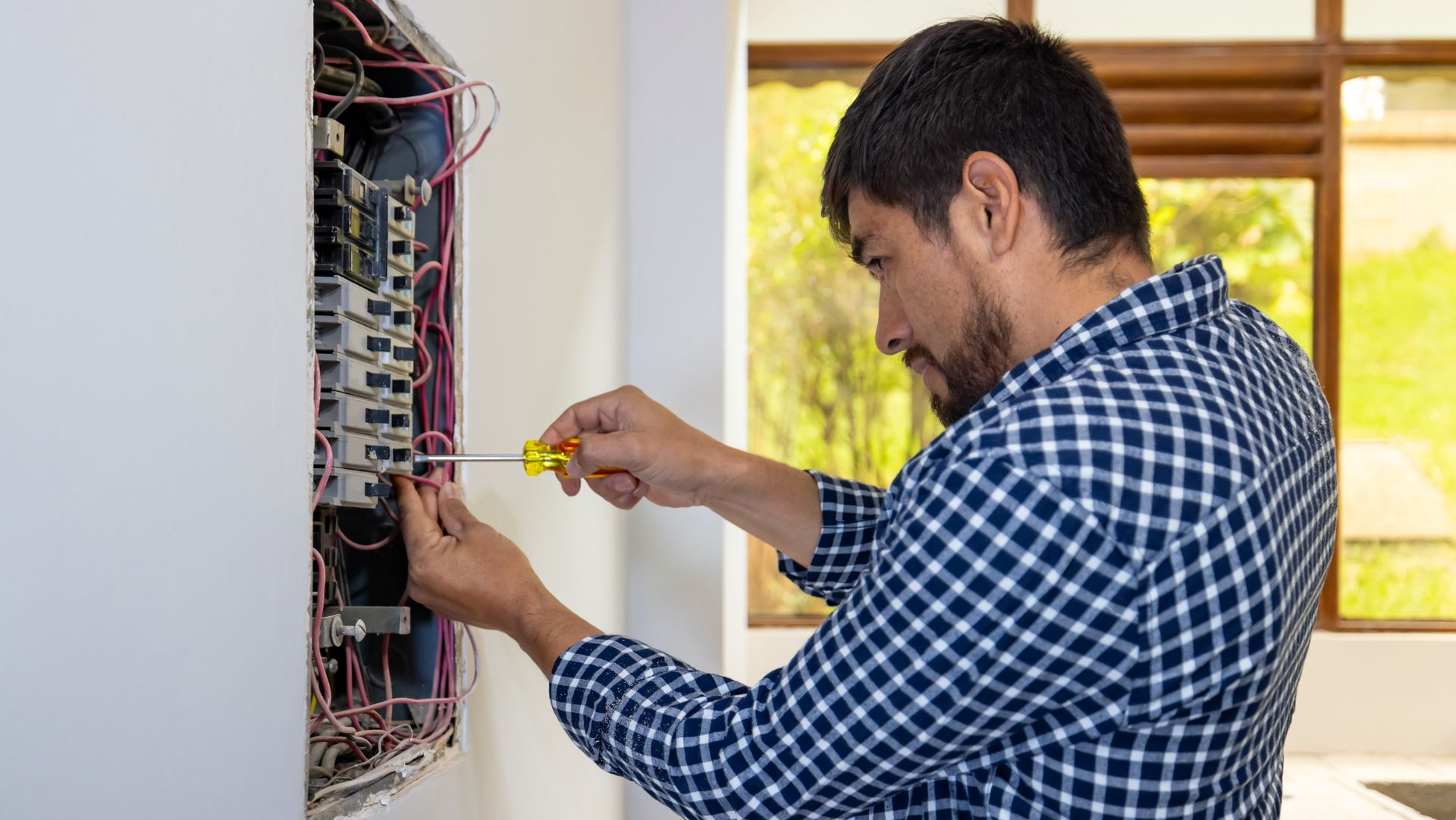 Man in plaid shirt, using a screwdriver on an open electrical panel mounted on a wall.