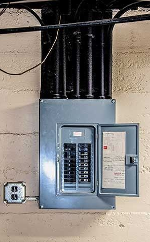 Electrical panel with open door, mounted on a concrete wall with conduit above and an outlet to the side.