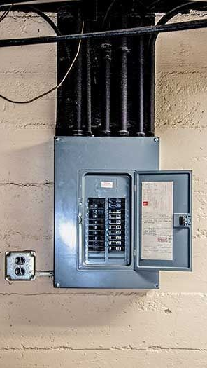 Electrical panel with open door, mounted on a concrete wall with conduit above and an outlet to the side.