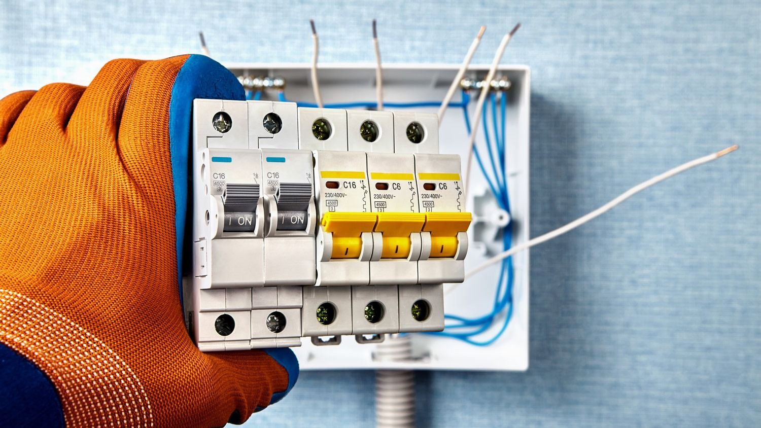 Hand holding a circuit breaker panel with exposed wires; orange glove.