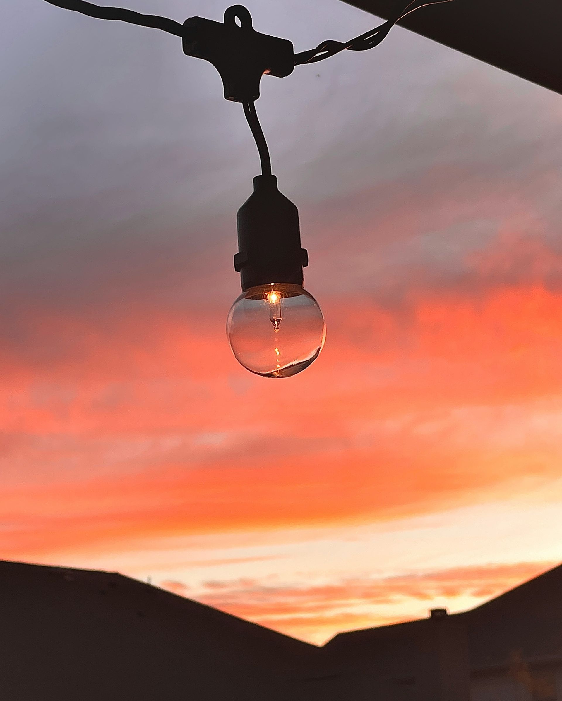 Lightbulb hanging against a vibrant orange and pink sunset sky with dark silhouettes of houses.