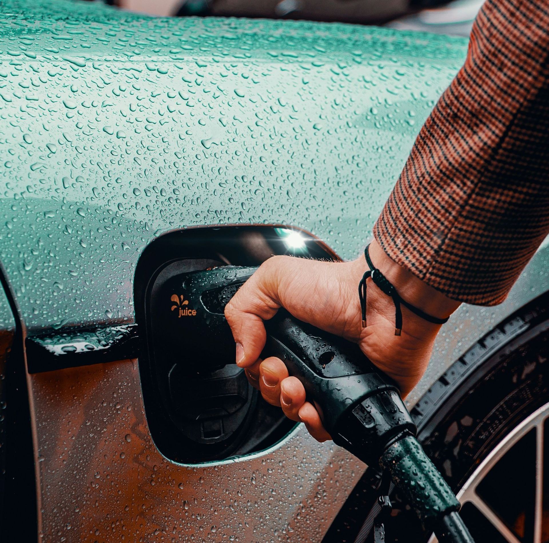 Person plugging a black charging cable into the port of a silver electric car in the rain.