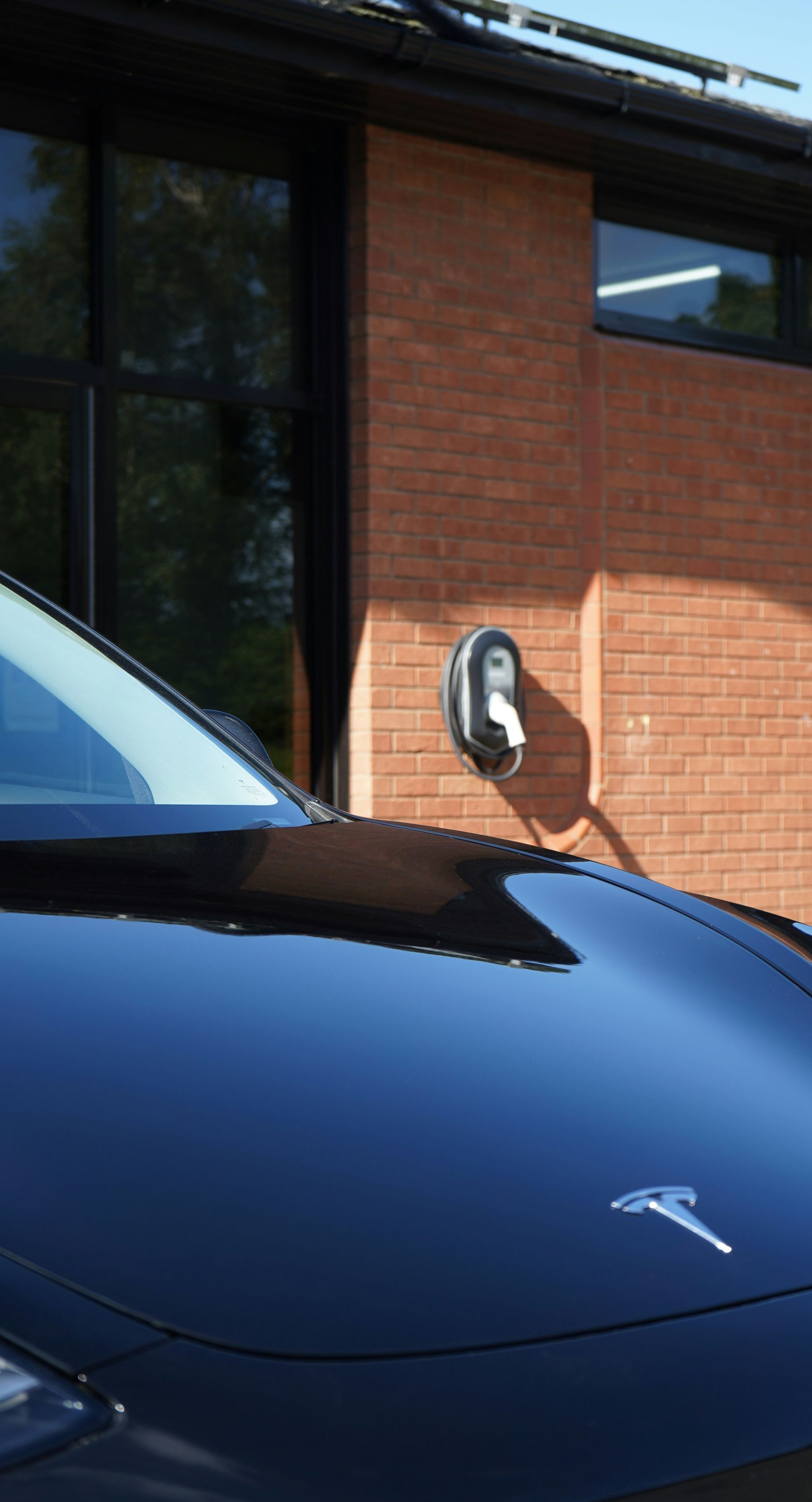 A black Tesla electric car charging at a home charging station mounted on a red brick wall.