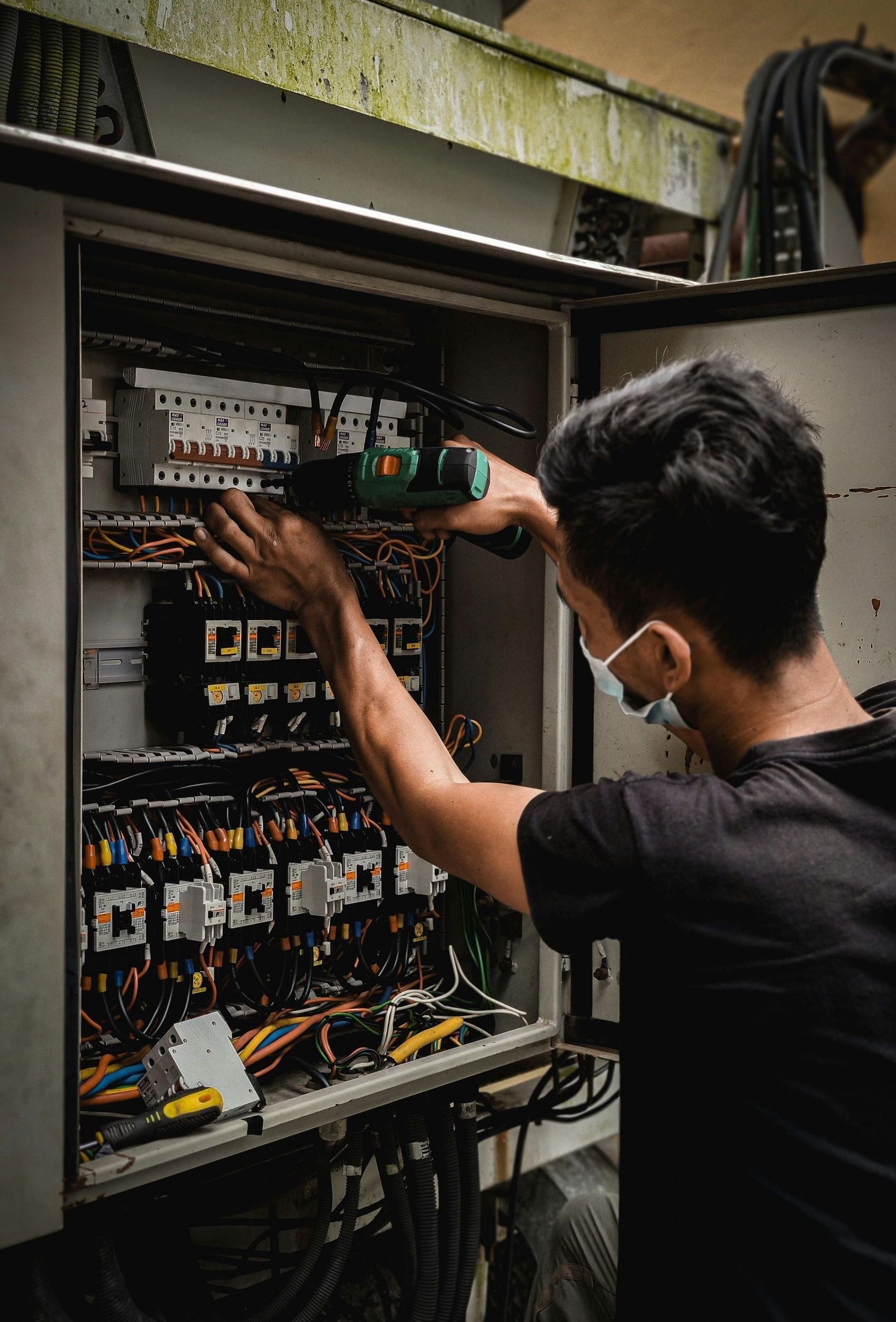 Man in mask using a drill to work on electrical wiring inside a gray cabinet.