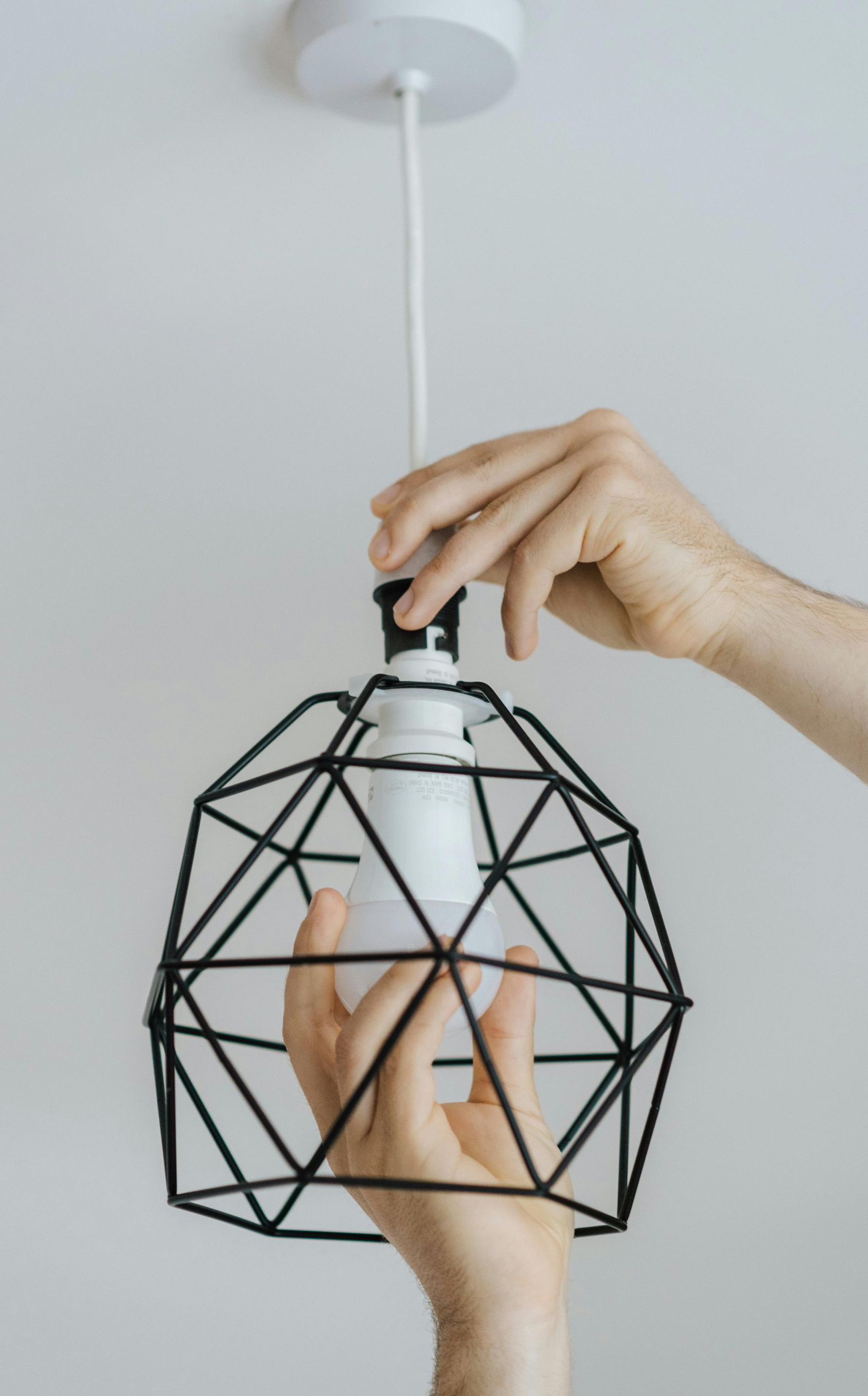 Person replacing a lightbulb in a geometric black wireframe pendant lamp against a white ceiling.