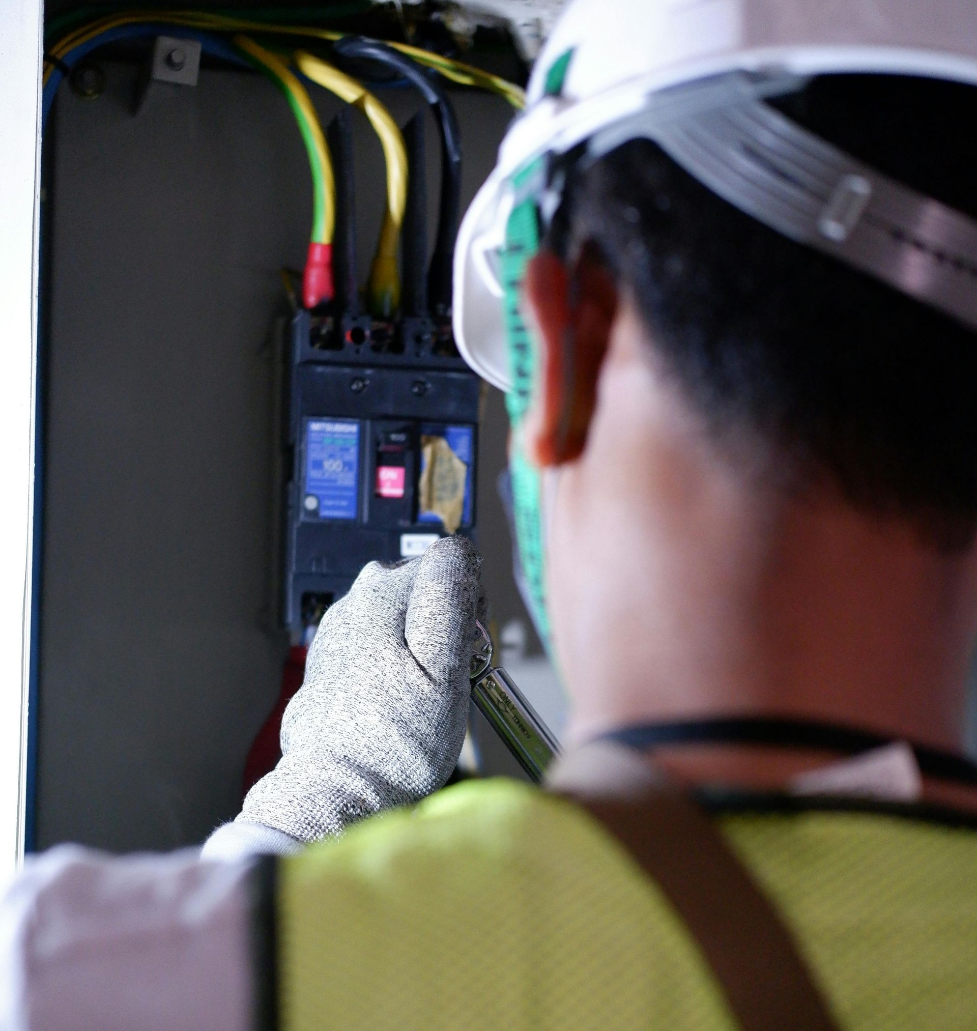 Electrician in hard hat and gloves, working on circuit breaker panel with yellow, green, and black wires.