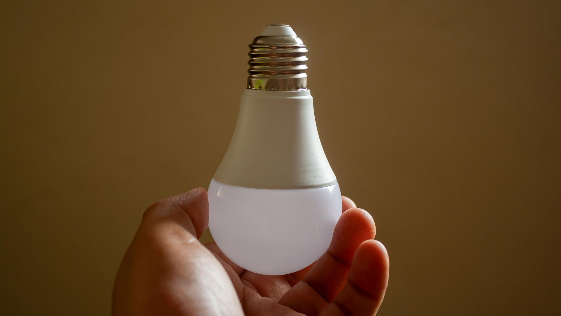 Hand holding a white LED lightbulb against a tan background.