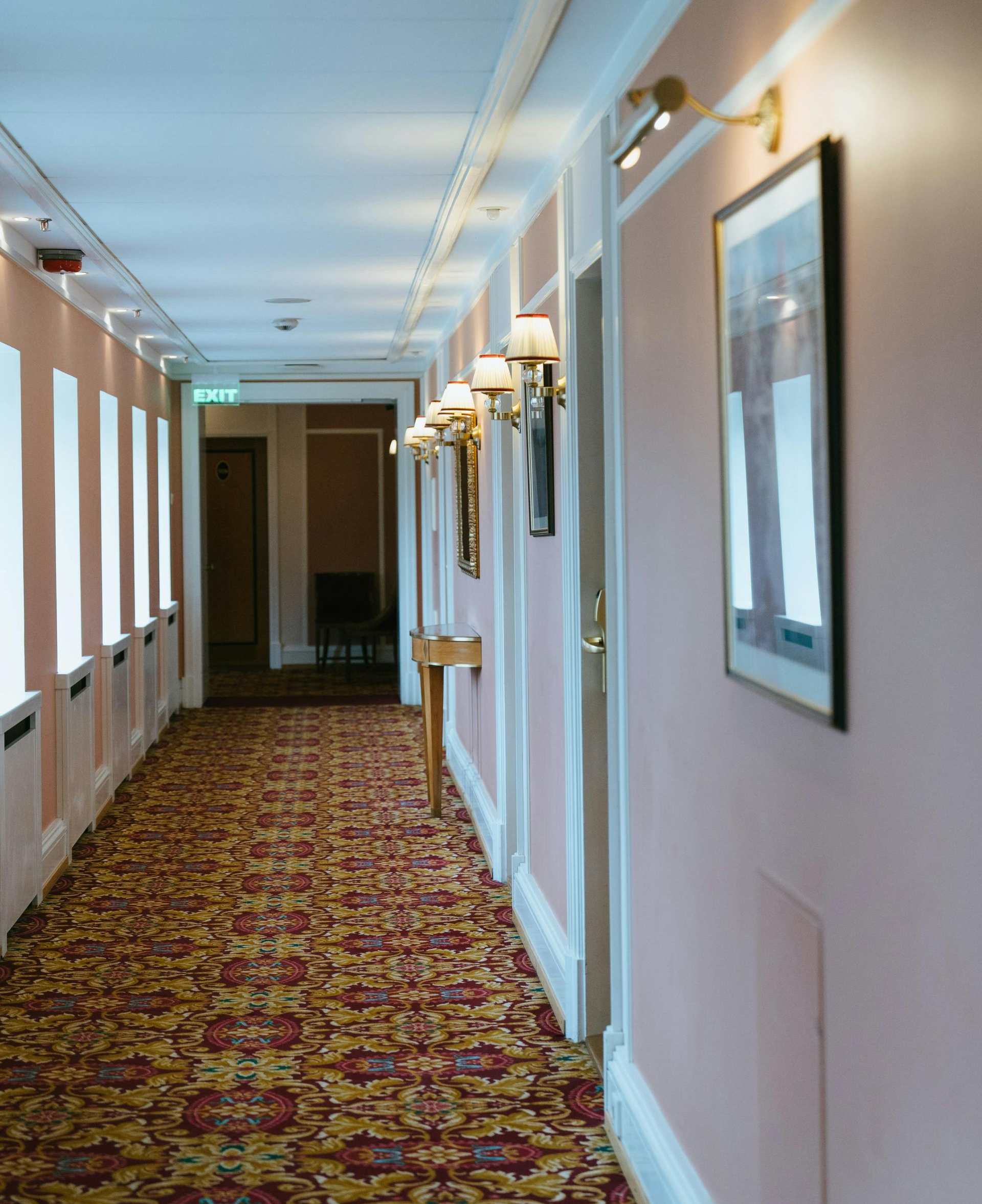 Hotel hallway with patterned carpet, pink walls, sconce lighting, and framed artwork.