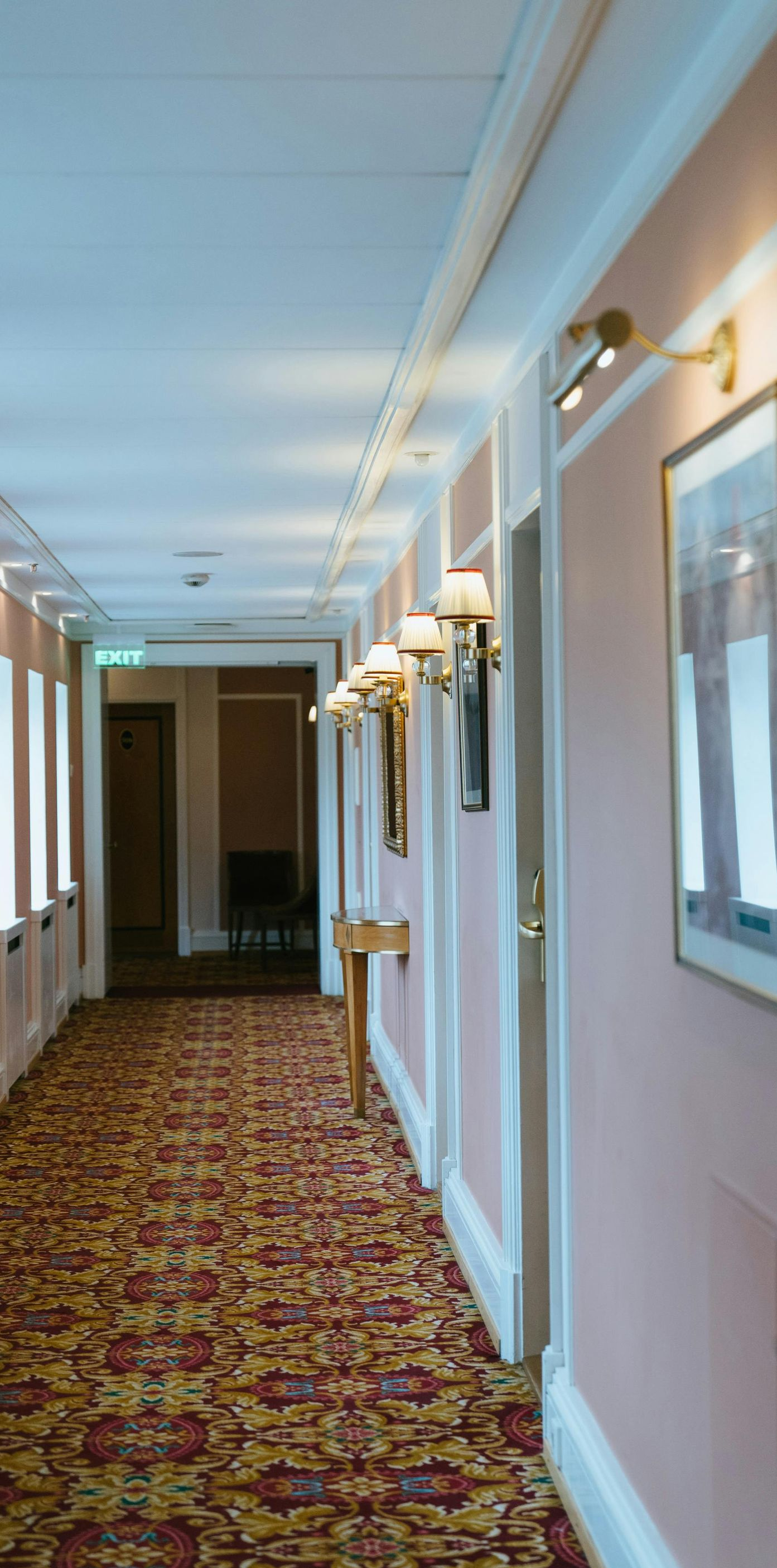 Hotel hallway with pink walls, patterned carpet, and sconce lighting.