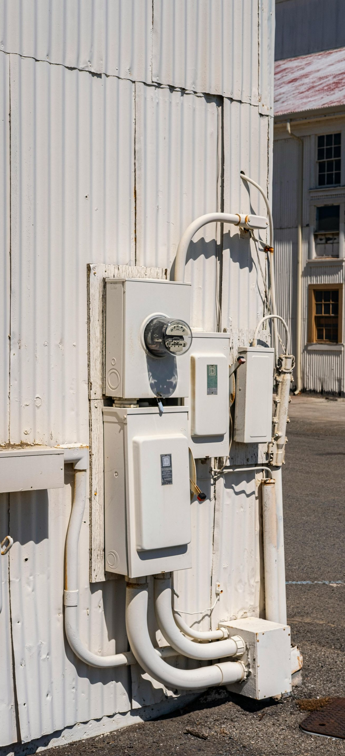 Electrical panel boxes on a white building's exterior. White conduit pipes connect the boxes. A second building is in the background.