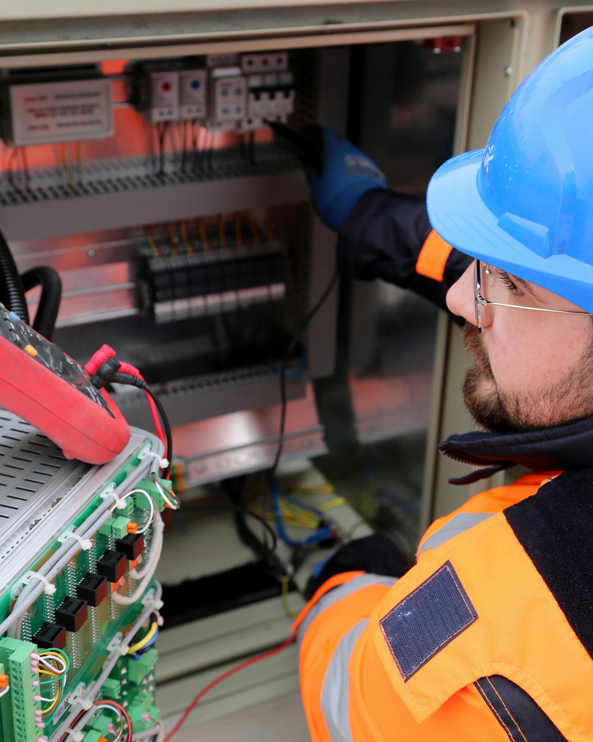Electrician in blue hard hat tests electrical panel with multimeter. Orange safety vest and gloves.