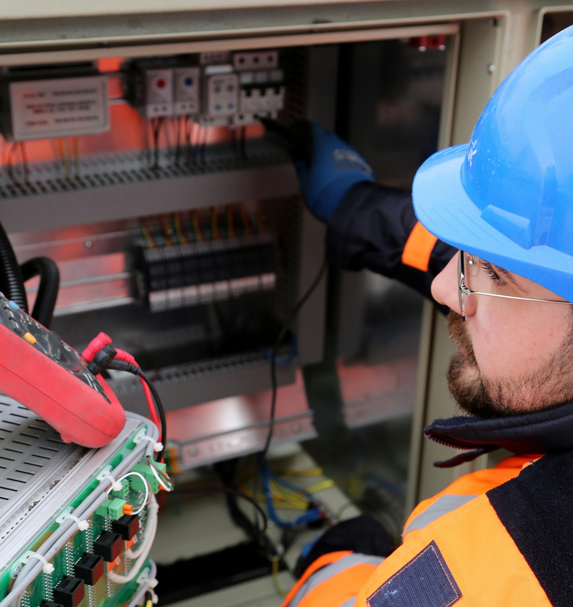 Electrician in a safety vest and hard hat, working on a control panel with a multimeter.