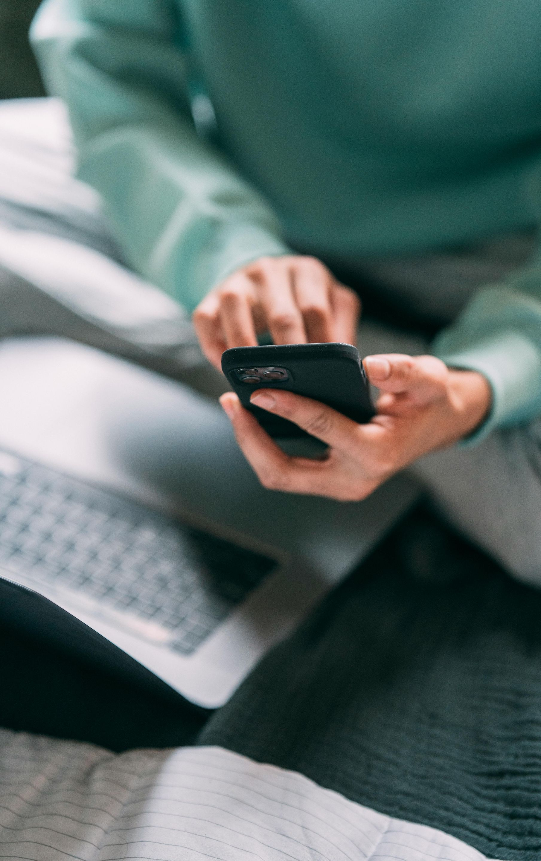 A person in a teal sweater sits on a bed, using a smartphone while a laptop rests nearby.