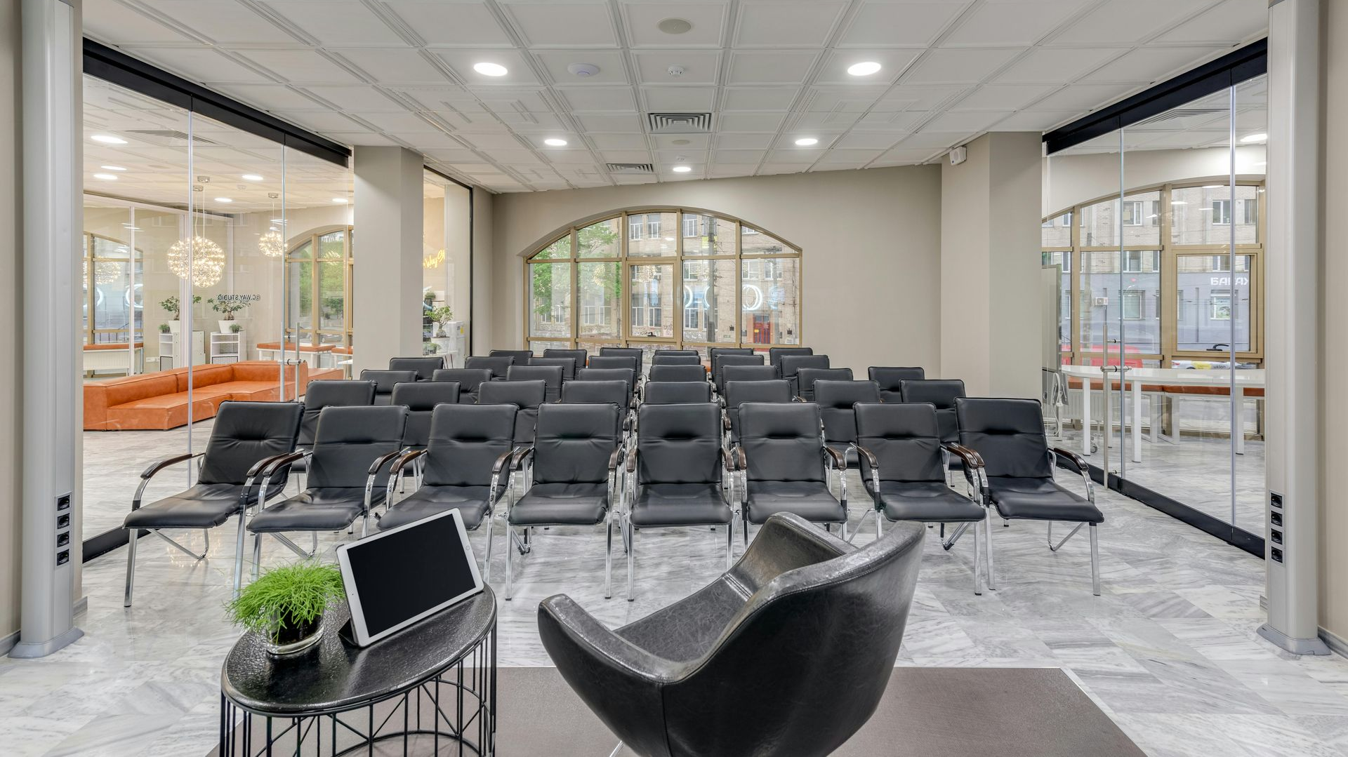 A conference room with rows of black chairs, a speaker's chair, and a small table with a tablet and plant.