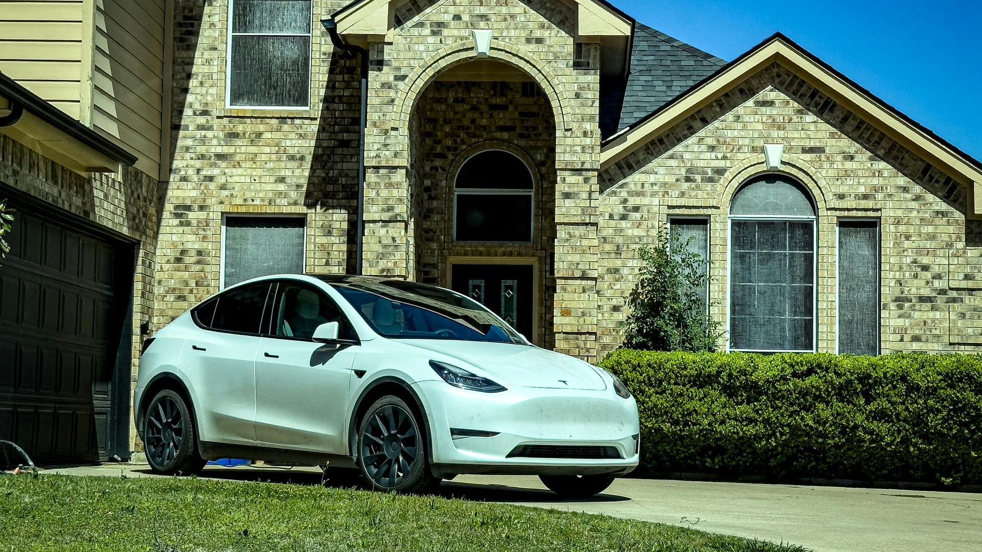 White Tesla Model Y parked in front of a two-story brick house on a sunny day.