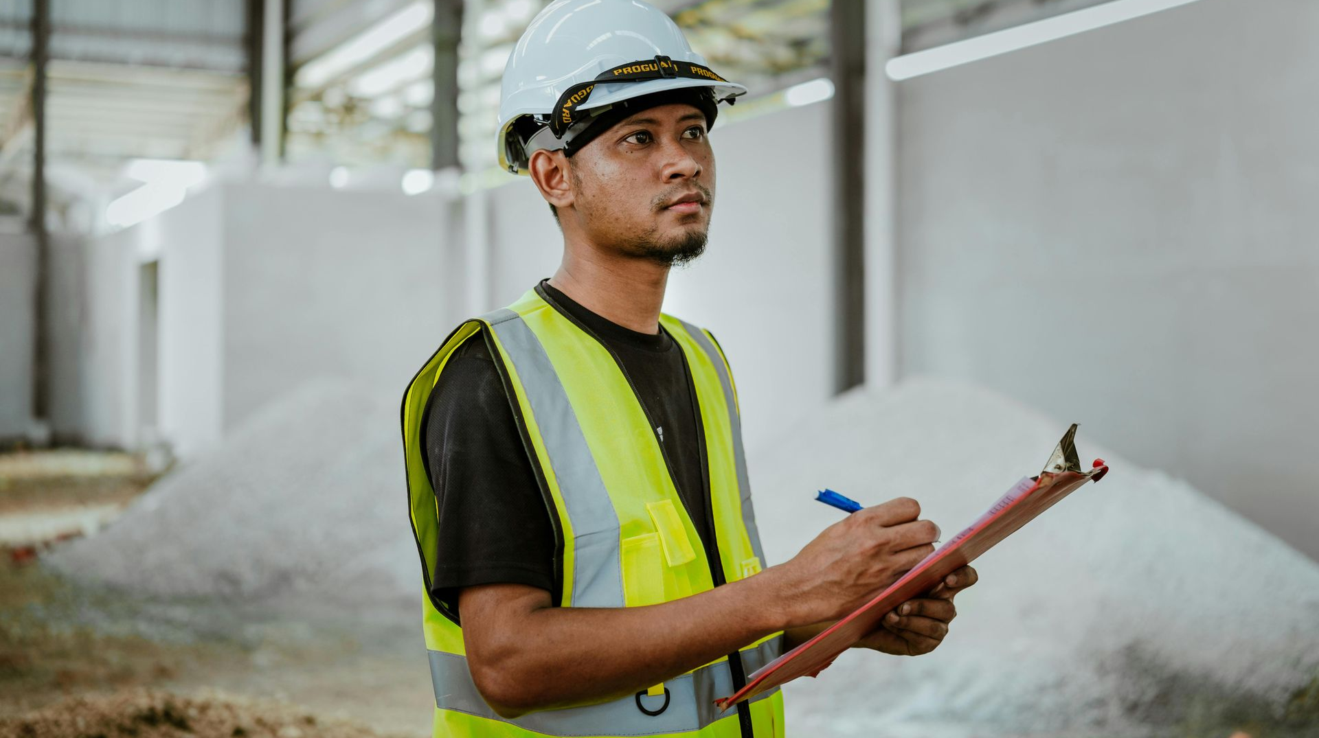 Construction worker in hard hat and safety vest, holding clipboard, inside a warehouse.
