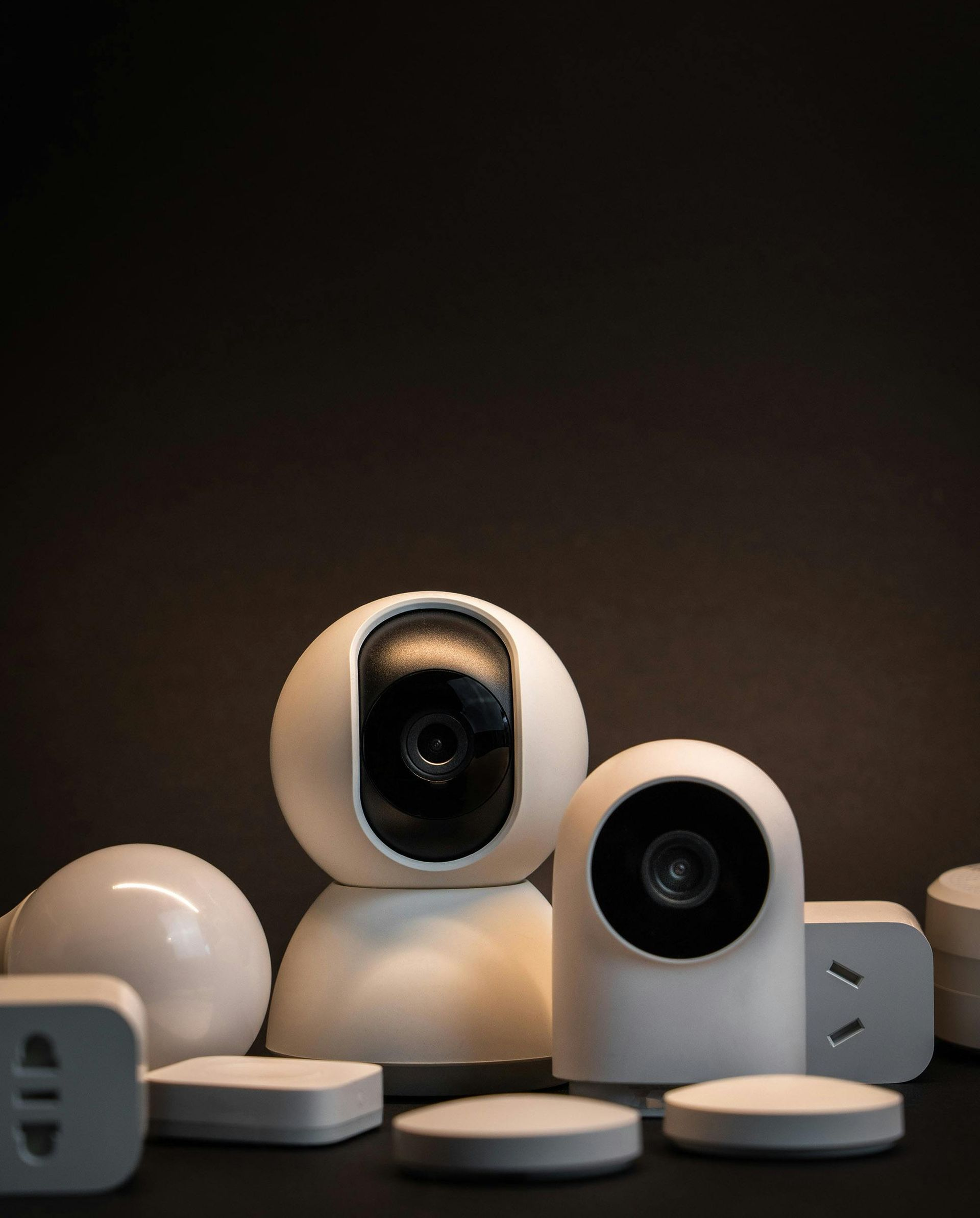 Two white smart home security cameras surrounded by various small smart home sensors and wall adapters on a dark surface.