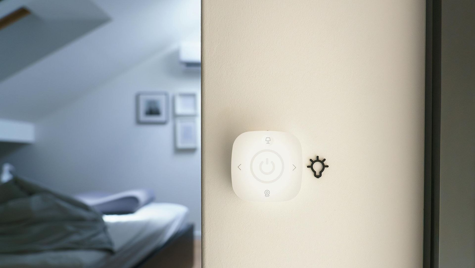 A wireless, square smart light switch mounted on a light-colored wall, with a partially visible bedroom in the background.