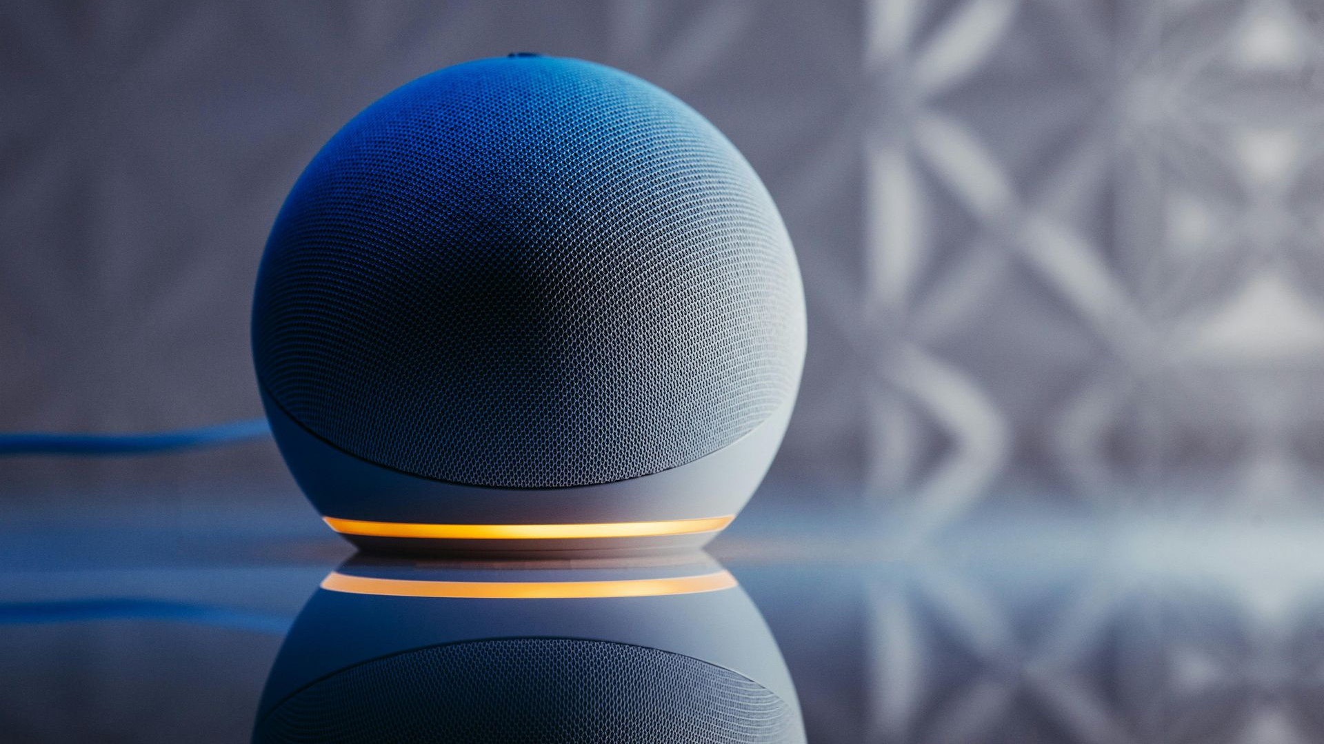 A blue spherical smart speaker sits on a reflective surface, illuminated by a glowing yellow ring light at its base.