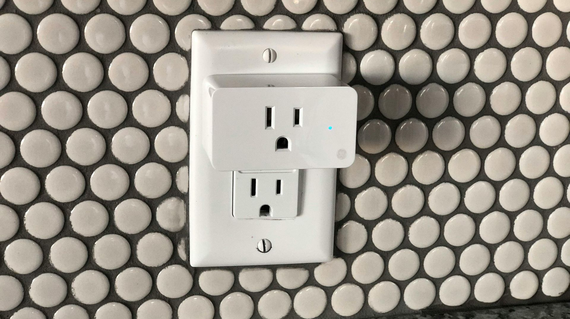 A white electrical outlet adapter is plugged into the top socket of a wall outlet against a white penny tile backsplash.
