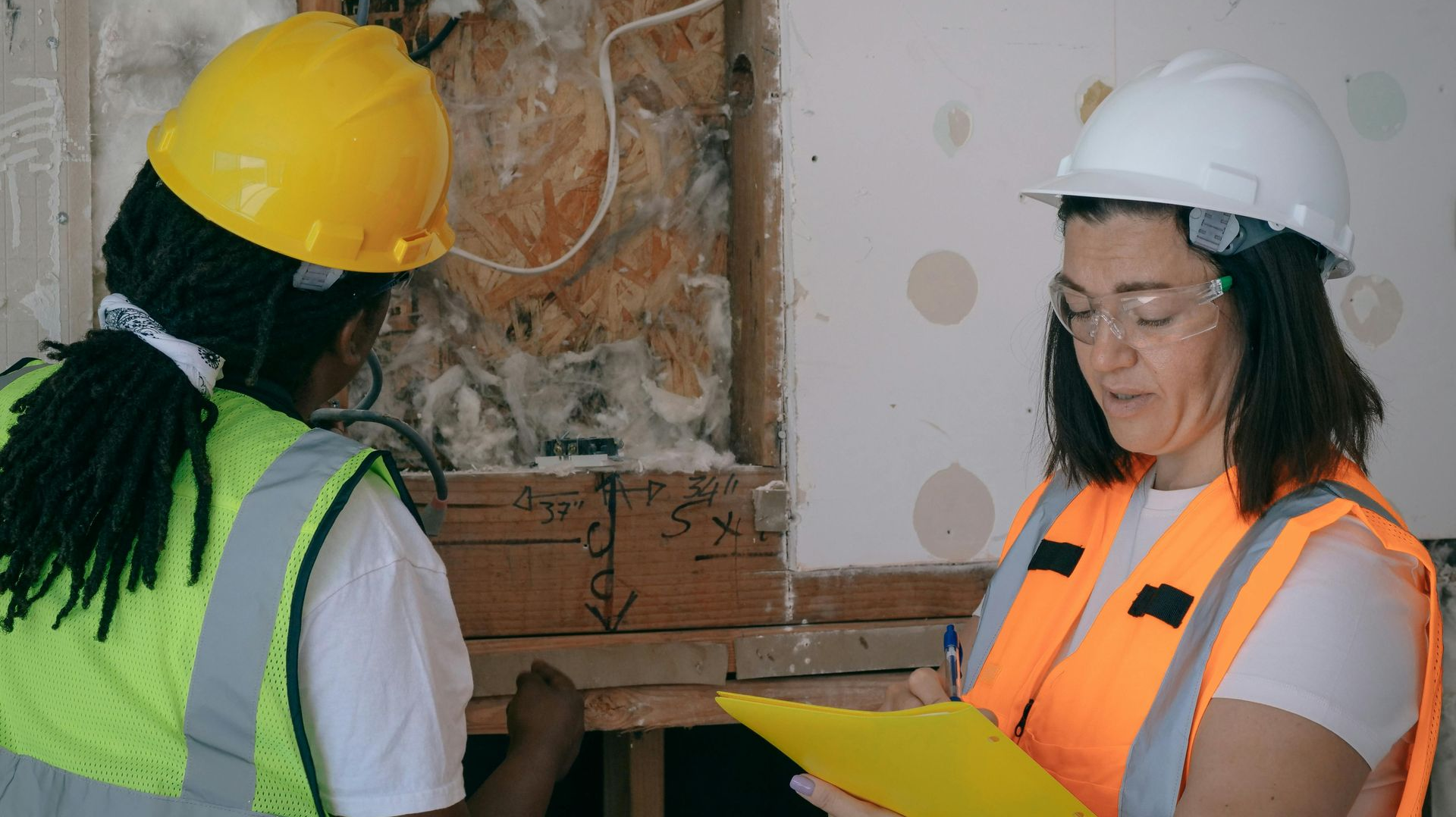 Two women wearing hard hats and vests inspect construction, one takes notes on a clipboard.