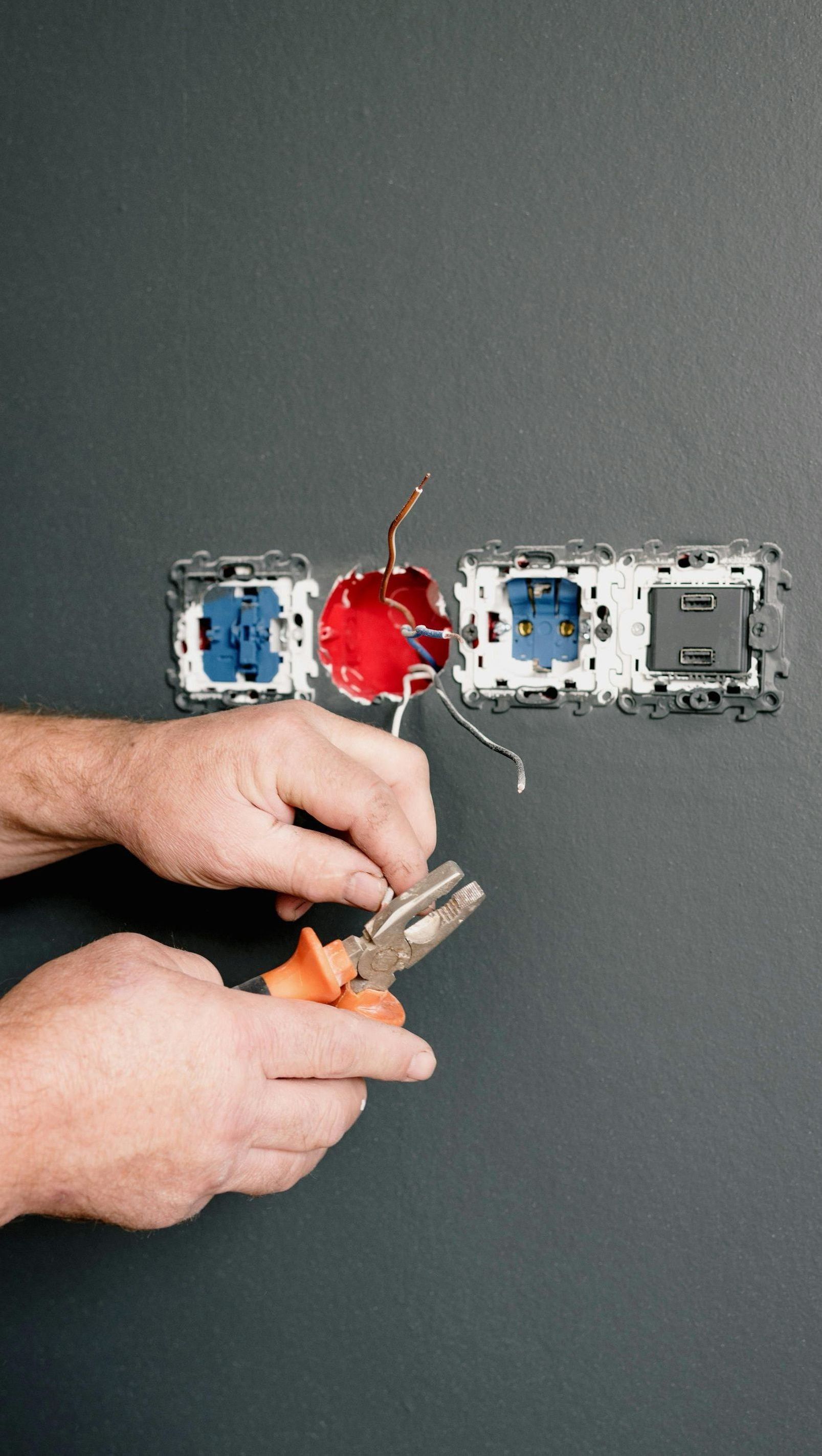 Hands wiring electrical outlet. Gray wall, orange pliers, exposed wires, and blue, red, and gray outlet components.