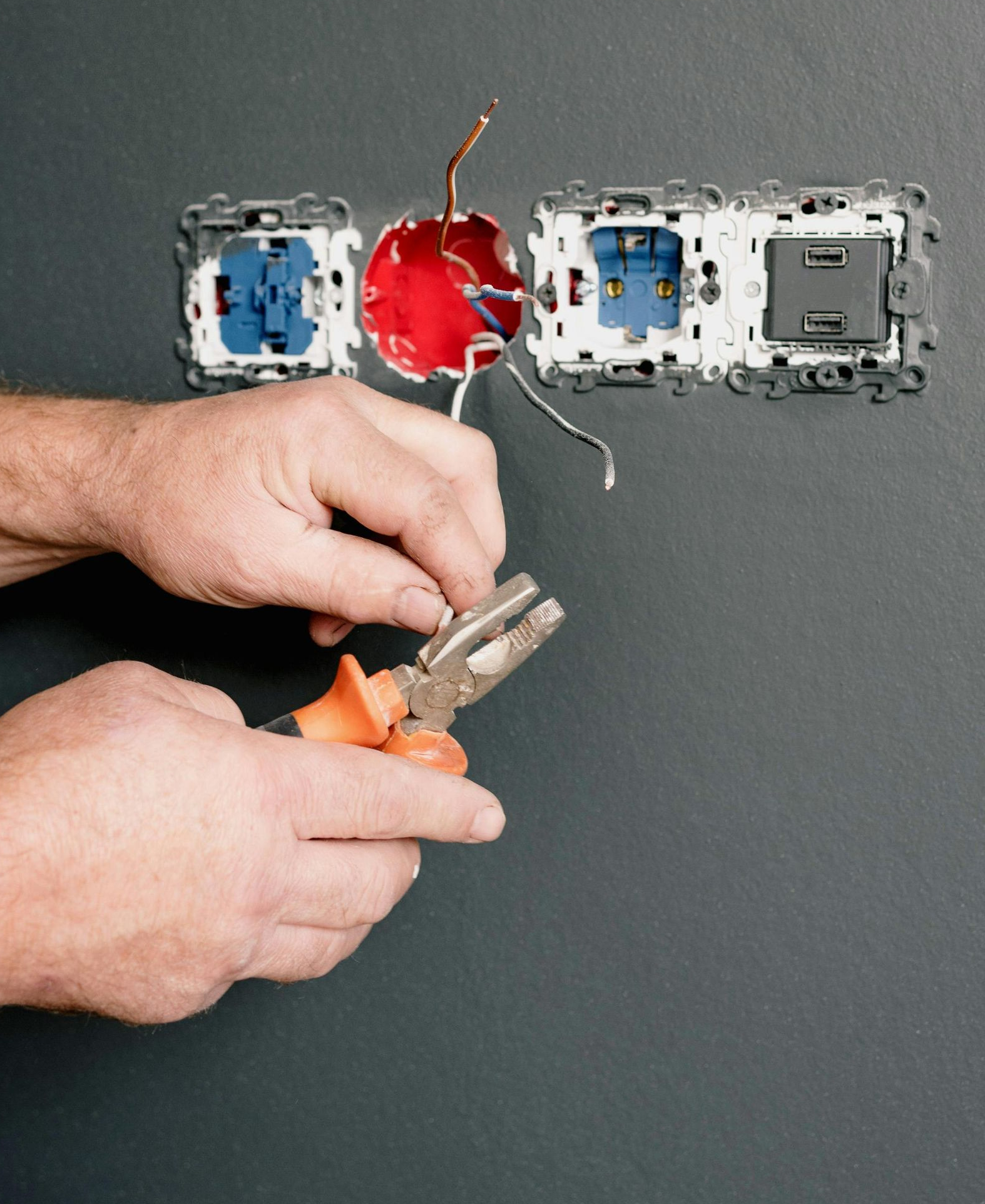 Person using pliers to work on electrical wiring in a wall outlet, with wires exposed.