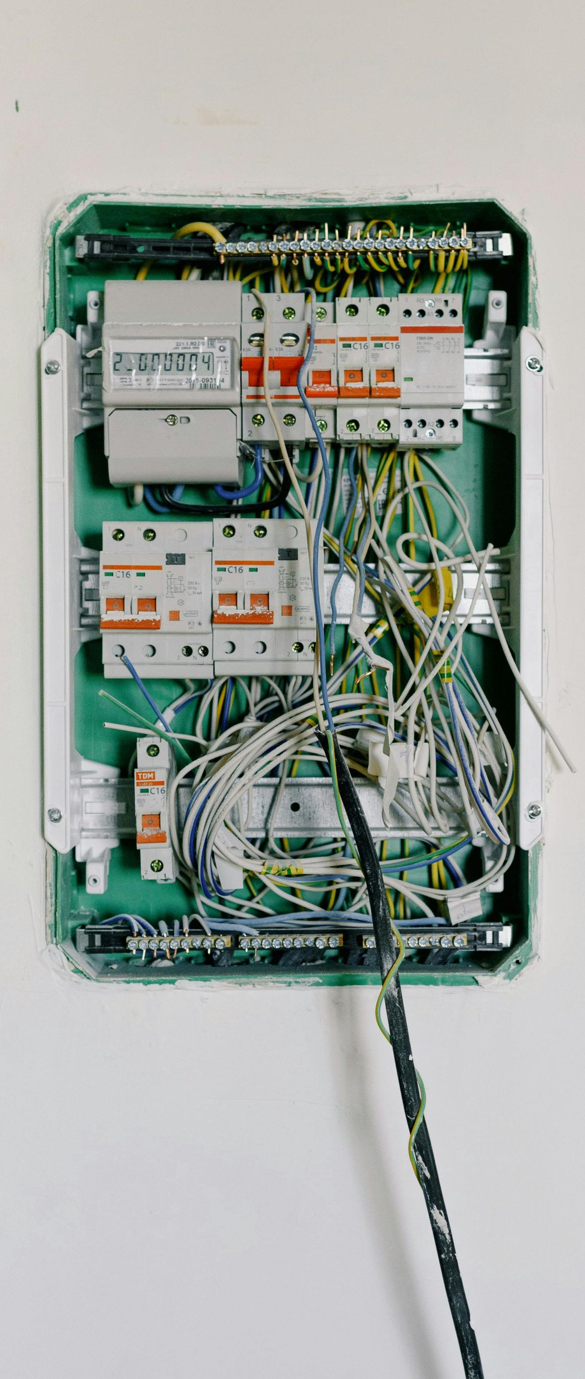 Open electrical panel with numerous wires and circuit breakers. Green box mounted on a white wall.