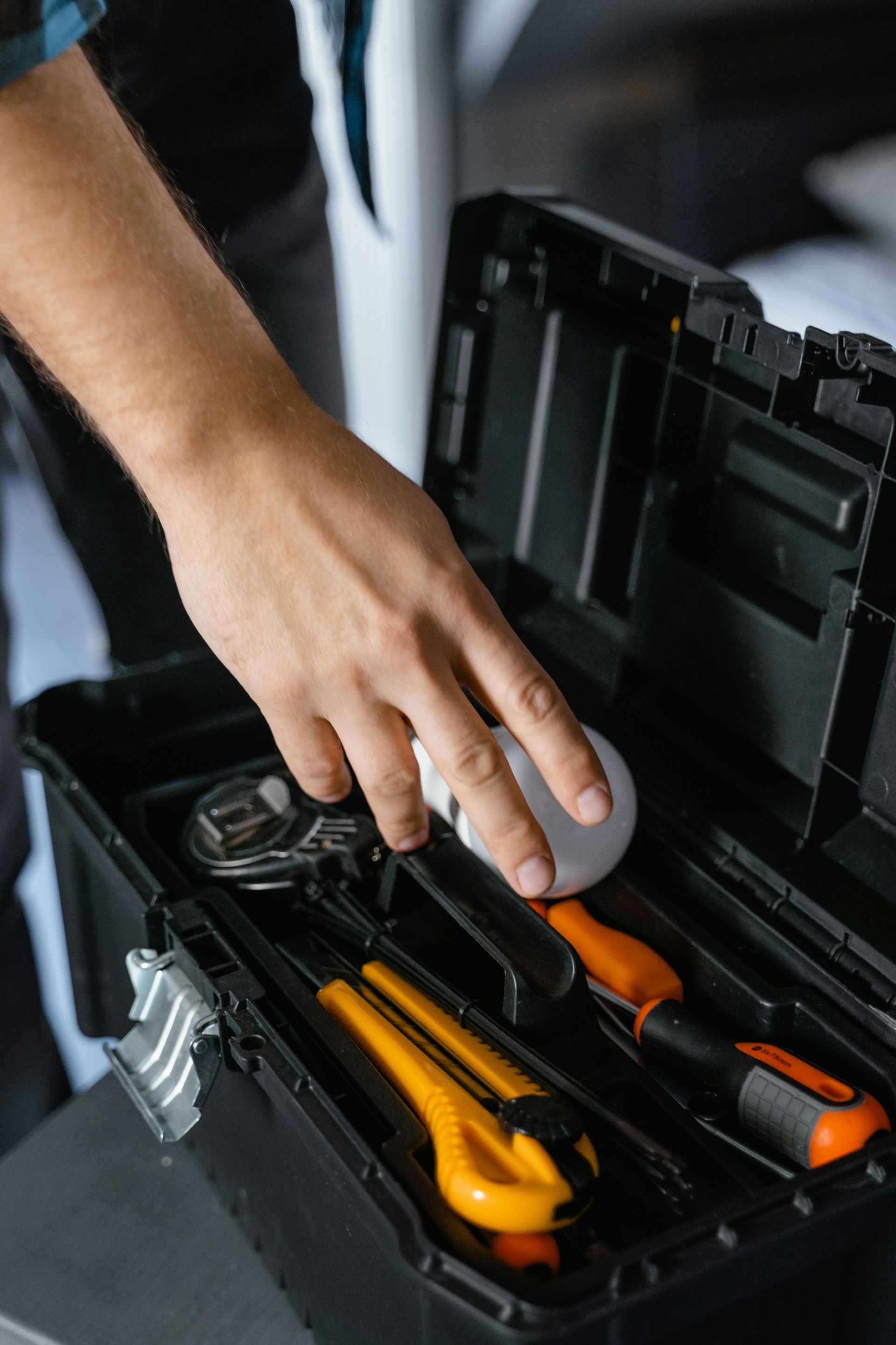 Person's hand reaching into a black toolbox filled with tools, including a wrench and screwdriver.