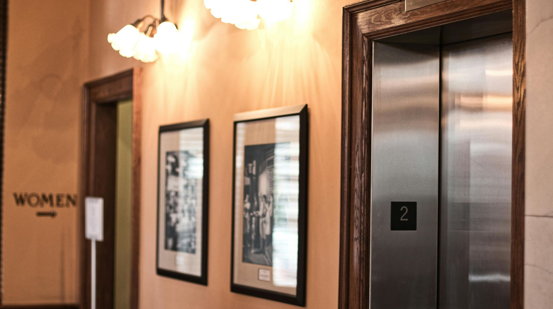Hallway with elevator, framed photos, and women's restroom sign on a peach-colored wall, with a light fixture overhead.