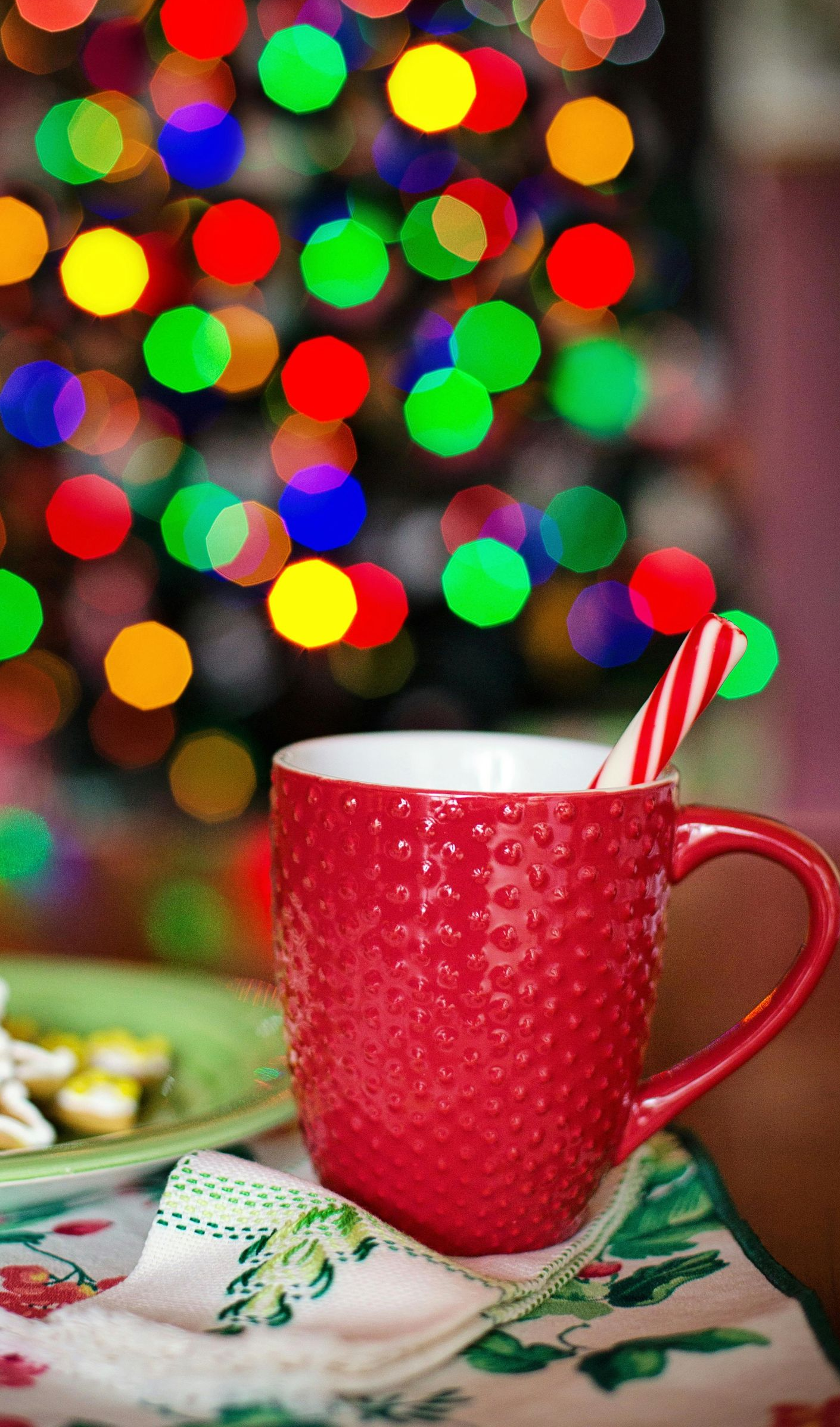 Red mug with candy cane on festive cloth, blurred Christmas lights in the background.