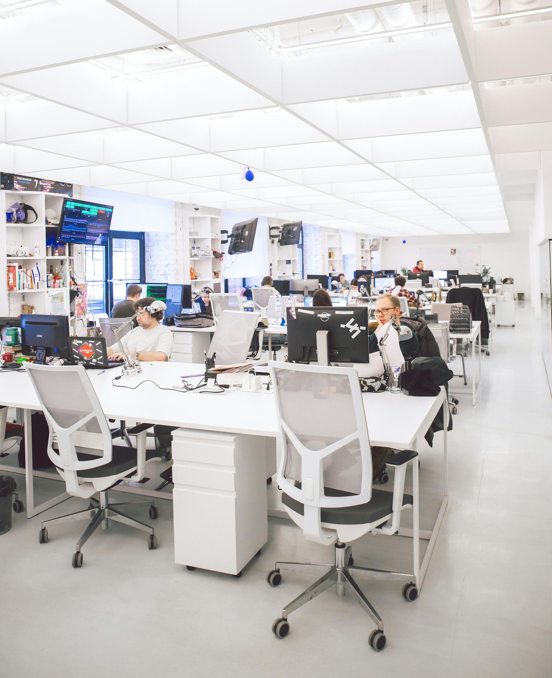 Bright office with white desks, chairs, and a large ceiling. People work on computers.