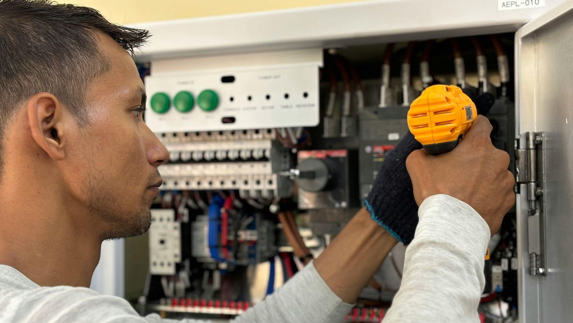 Man in grey shirt working on an electrical panel, holding a yellow tool.