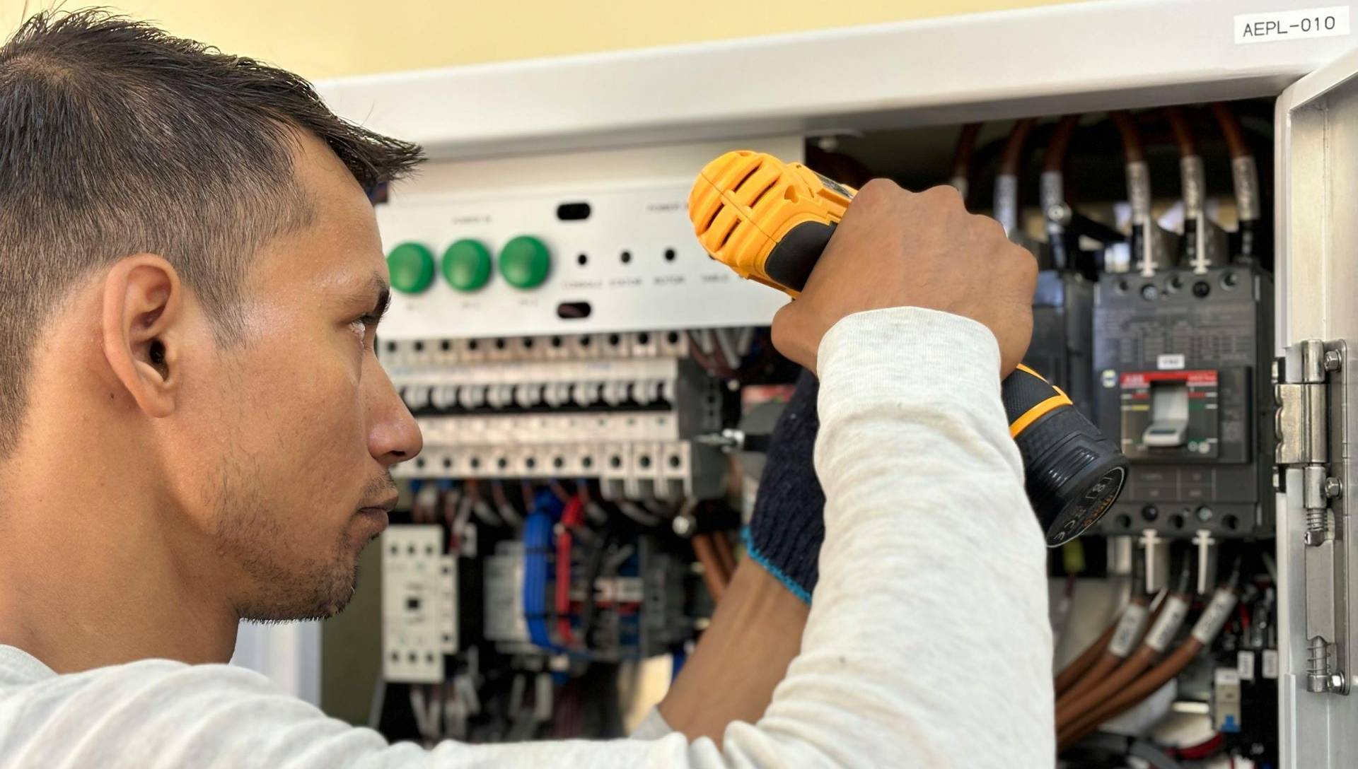 Electrician using a power drill on electrical panel, wearing gloves, indoors.