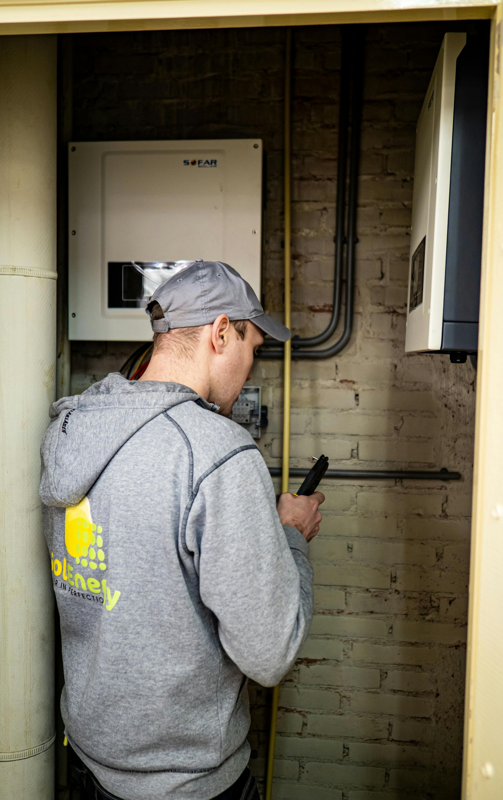 Man in gray hoodie examines electrical equipment in a brick-walled room.