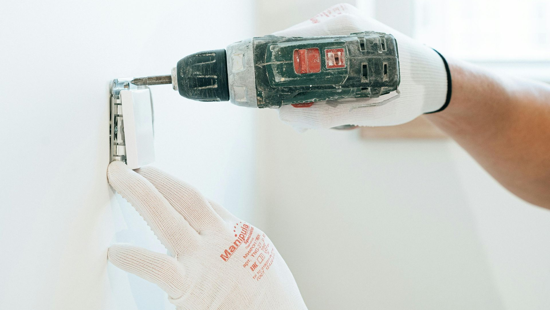 Person wearing white gloves, using a drill to install an electrical outlet on a white wall.