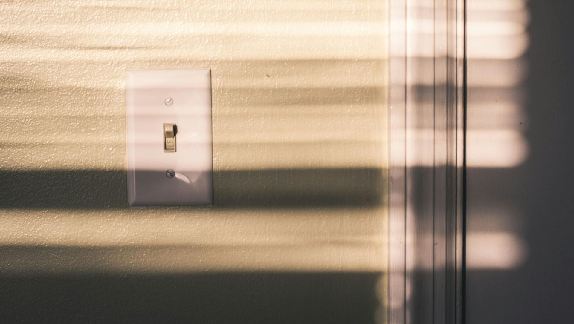 Light switch on a wall, with sunlight and shadows from blinds.