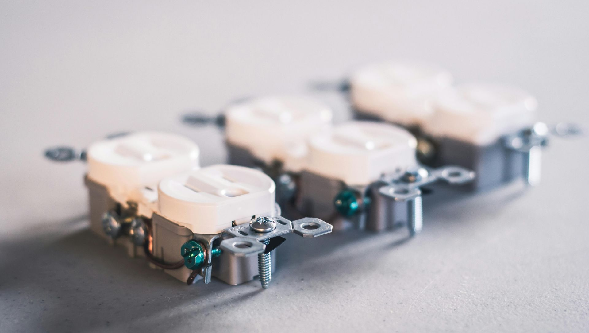 Three electrical duplex outlets, white receptacles with silver metal frames, on a light gray surface.