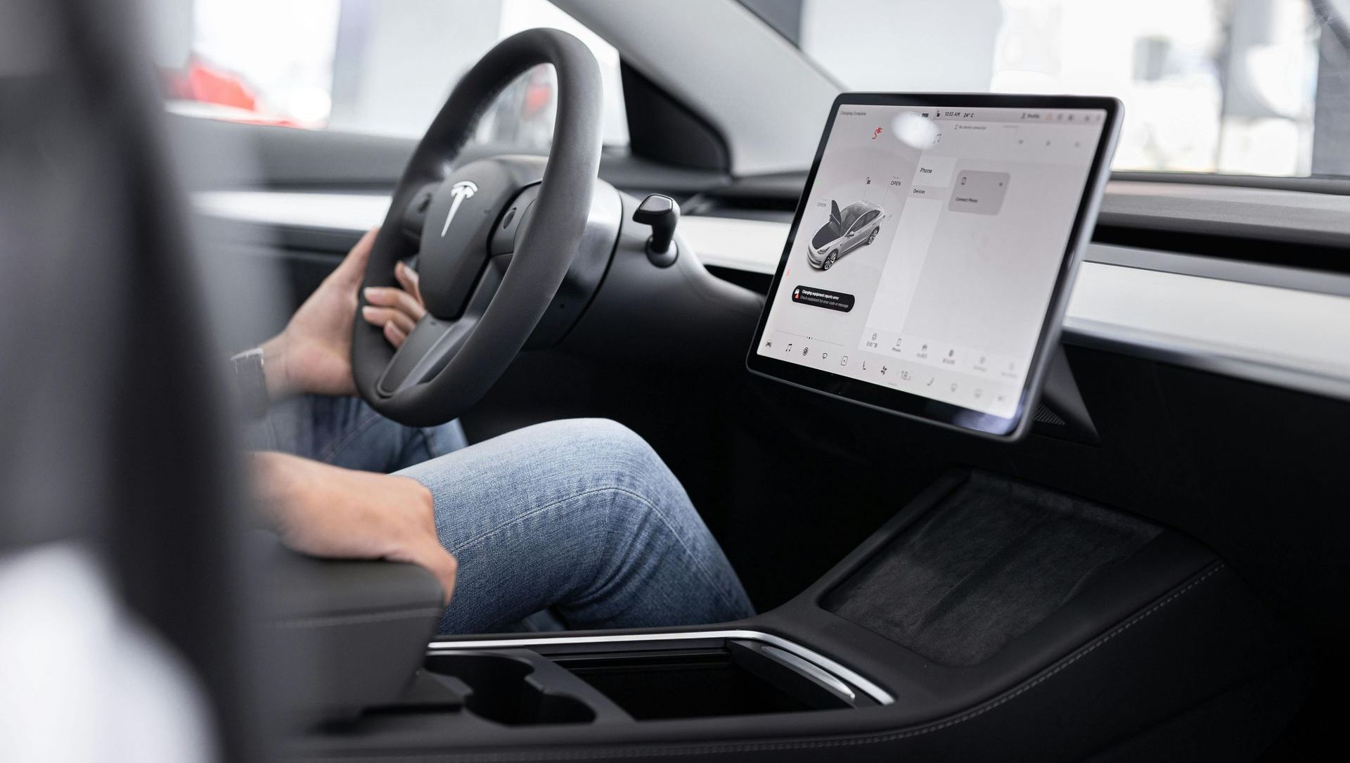 Inside a Tesla car, a person grips the steering wheel. A large touchscreen display is visible on the dashboard.