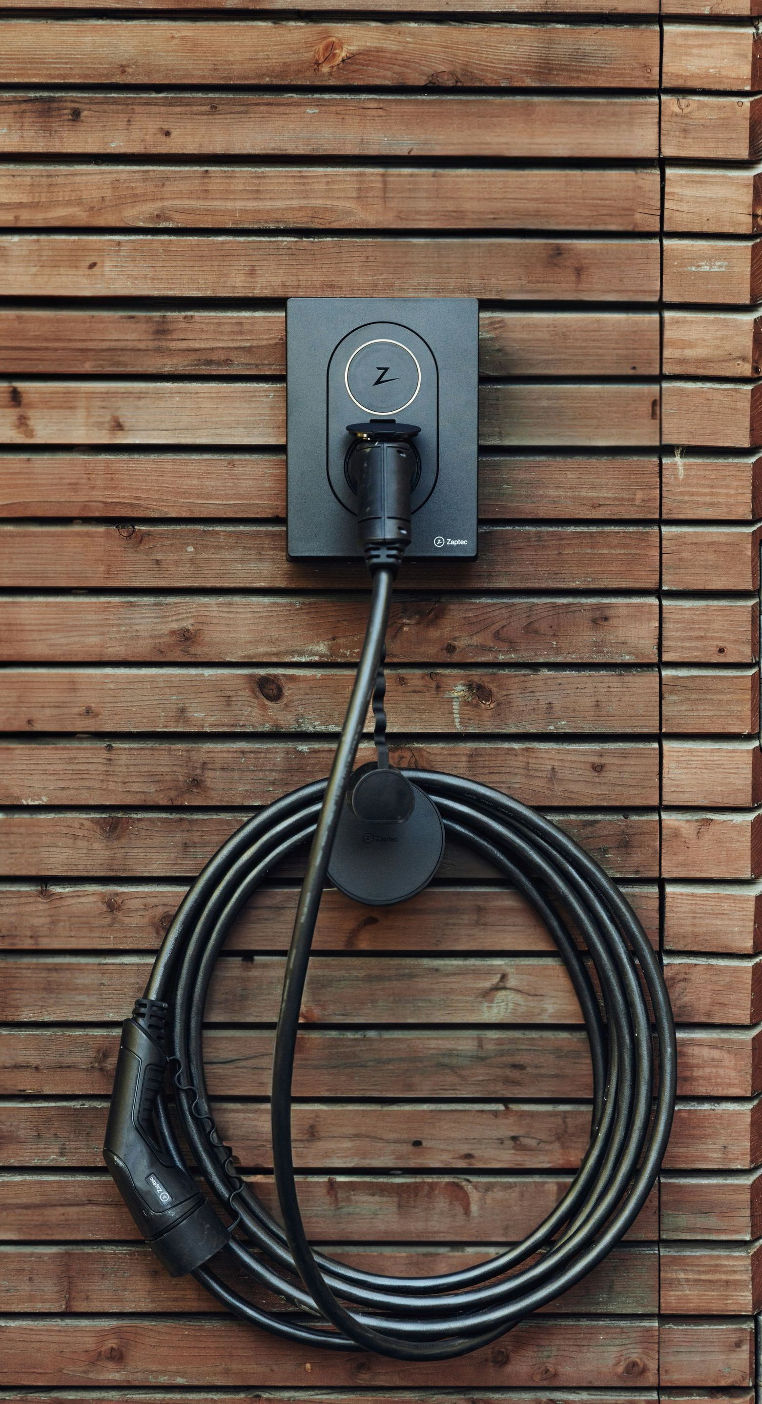An EV charging station mounted on a wood-paneled wall. The black cable is coiled and plugged into the charger.